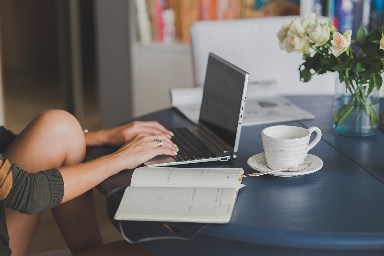 women, writing on laptop, notebook open, coffee, blogging